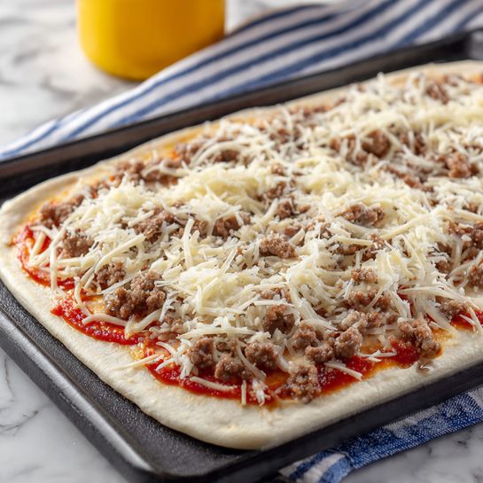 A close-up view of a raw rectangular pizza on a black tray, placed on a white marbled surface. The pizza has three visible layers: the bottom layer is a light beige dough, spread thin with some edges showing, with red tomato sauce dotted around the crust edges. The middle layer is a generous spread of shredded white cheese scattered evenly across the surface. On top, there are small brown pieces of cooked ground meat distributed sparsely but covering most of the cheese. The background shows a striped blue and white cloth and a yellow-labeled bottle blurred out. Photo taken with an iphone --ar 4:5 --v 7