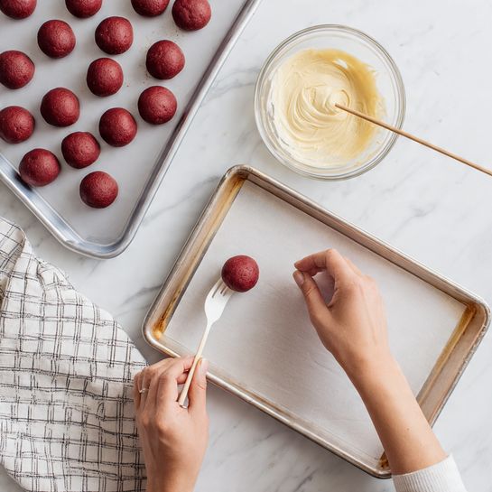The image shows two white coated round balls placed on a parchment-lined silver baking tray on a white marbled surface. One ball is on the tray, fully coated with smooth white texture, while the other is being held on a white fork by a woman's hand and dipped carefully with a wooden stick from another woman's hand. To the top left, several red dough balls are arranged on another silver baking tray lined with parchment paper, and a clear glass bowl filled with white creamy mixture is also present near the top center. A white and black checkered cloth is partially visible on the bottom left. photo taken with an iphone --ar 4:5 --v 7
