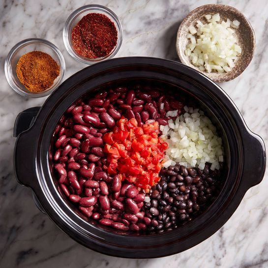 A top view of a black slow cooker filled with three types of beans: dark red kidney beans, lighter red beans, and black beans forming a textured base layer. On top of the beans in the center, there is a small pile of finely chopped bright red tomatoes. To the left side, there is a layer of finely chopped white onions. Two small glass bowls positioned near the edges hold reddish-brown spice powders. The slow cooker rests on a white marbled textured surface. photo taken with an iphone --ar 4:5 --v 7