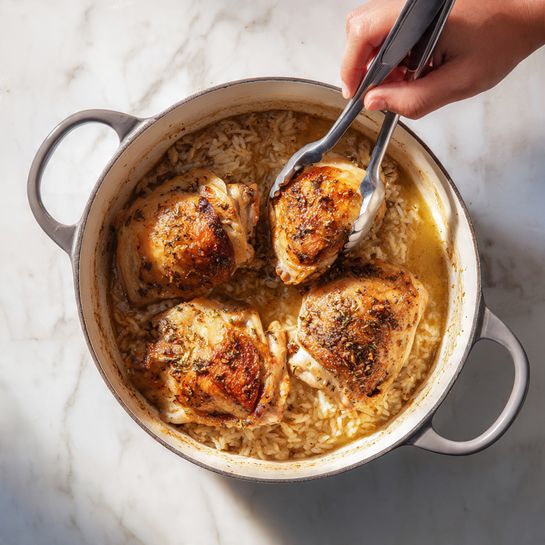 The image shows a round white pot with gray handles on a white marbled surface. Inside the pot are three cooked seasoned chicken pieces with a brown and slightly crispy texture, resting on the bottom. In the left side of the image, a woman's hand is using metal tongs to hold one chicken piece. On the right side of the image, the same three chicken pieces sit on a layer of light brown rice soaking in a broth that covers part of the rice and surrounds the chicken. The rice looks soft and creamy, with a slight shine from the liquid. The pot and food are well-lit with natural light showing clear textures and colors. photo taken with an iphone --ar 4:5 --v 7