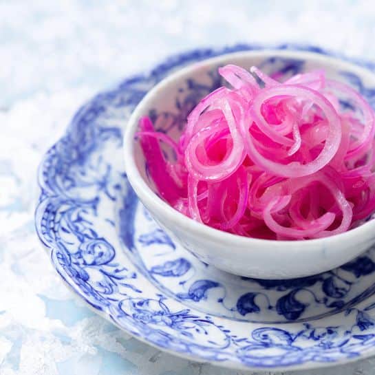 A small white bowl filled with thin, spiraled bright pink pickled onions, showing a slightly translucent and wet texture. The bowl is placed on a white plate decorated with intricate blue floral patterns. The background and surface have a white marbled texture, giving a clean and fresh look to the image. Photo taken with an iphone --ar 4:5 --v 7