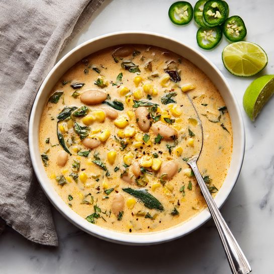 A creamy soup in a white bowl filled with layers of light orange broth mixed with white beans, bright yellow corn kernels, and small green herb pieces spread evenly throughout. There are small bits of darker charred corn adding texture on top. A metal spoon rests inside the bowl on the right side. The bowl is placed on a white marbled surface with lime wedges and green herbs arranged around it, along with sliced jalapeño peppers and a soft gray cloth on the left. Photo taken with an iphone --ar 4:5 --v 7