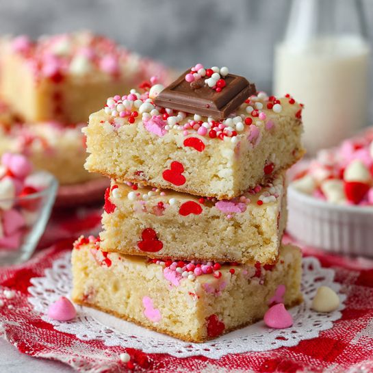 A stack of four square blondie bars sits on a white lace doily over a red and white checkered cloth, each bar light golden brown with small red candy pieces mixed inside. The top blondie has a heart-shaped milk chocolate piece in the center, covered with pink, red, and white round sprinkles. Pink, white, and red candy-coated chocolate pieces are embedded randomly across the top blondie, adding color pops. In the soft-focused background, there is a white bowl filled with pink, red, and white candy-coated chocolates and a white plate with more blondie bars. The whole scene rests on a white marbled texture. photo taken with an iphone --ar 4:5 --v 7