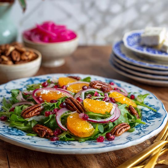 A white plate with blue floral patterns holds a fresh salad with three clear layers: the base layer is green arugula leaves spread all over, the middle layer has bright orange mandarin slices and deep red pomegranate seeds scattered evenly, and the top layer features brown pecan nuts and thin rings of pink pickled onions arranged lightly. In the background, three small white bowls hold pink pickled onions, white cheese, and more pecans, slightly blurred. To the right, a stack of blue and white patterned plates and three golden forks lie on a wooden table with a white marbled texture background. The photo taken with an iphone --ar 4:5 --v 7