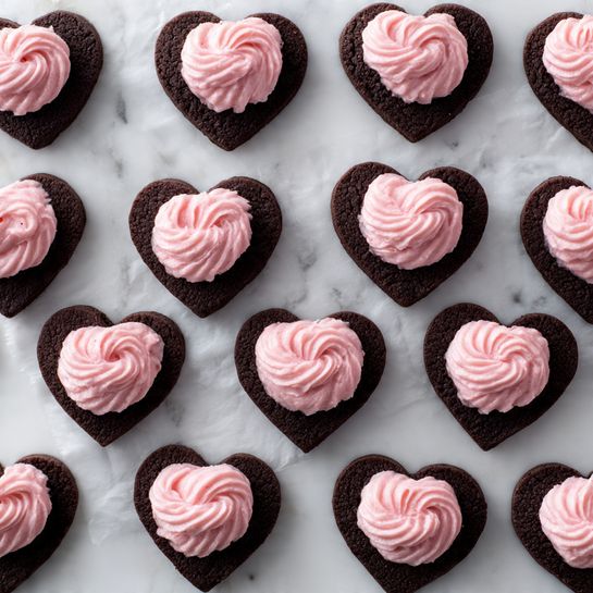 The image shows a neat arrangement of 30 small heart-shaped chocolate cookies on white parchment paper, placed on a white marbled surface. The cookies are laid out in 6 rows and 5 columns, alternating between plain dark chocolate hearts and ones topped with a dollop of smooth, pink cream frosting. The frosting has a soft, swirled texture and is centered on top of every other cookie, creating a pattern of dark brown and pink hearts. The overall look is clean and organized with a clear contrast between the dark brown cookies and bright pink frosting. Photo taken with an iphone --ar 4:5 --v 7