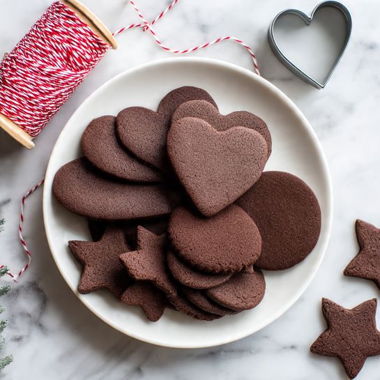 A white round plate holds three types of chocolate cookies arranged in layers: large heart-shaped cookies on the top layer, medium circular cookies on the right side overlapping slightly, and star-shaped cookies on the bottom left side forming a small pile. The cookies are dark brown and smooth-textured. Some cookies also lie scattered on the white marbled surface around the plate. Nearby, there is a spool of red and white twisted string with some string loosely laid across the top part of the image, and a light gray, heart-shaped cookie cutter sits near the spool. Photo taken with an iphone --ar 4:5 --v 7
