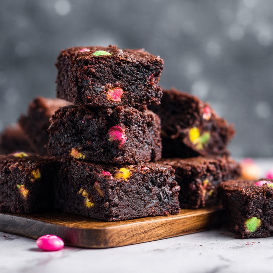 A stack of square chocolate brownies sits on a dark wooden tray against a dark blurred background, each brownie showing a rich, dense, and moist texture with a slightly cracked top layer. Scattered through the brownies are colorful candy pieces, mainly pink, yellow, and green, which add bright splashes of color and a slightly glossy texture. The brownies are arranged in a loose pile with some pieces overlapping, and a single pink candy lies beside the tray on the surface. The scene is set on a white marbled texture. Photo taken with an iphone --ar 4:5 --v 7
