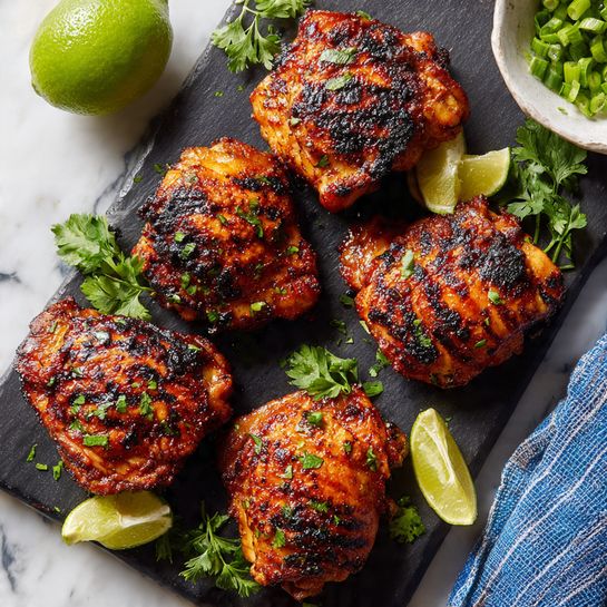Four pieces of grilled chicken with a dark brown charred outside are arranged on a black cutting board. There are bright green fresh parsley leaves placed around the chicken and four lime wedges, two on the top right and two on the bottom left. A whole lime rests on the top left corner of the board. A white bowl with chopped green onions is partially visible in the top right corner. A blue and white striped cloth is on the bottom right corner on a white marbled surface photo taken with an iphone --ar 4:5 --v 7