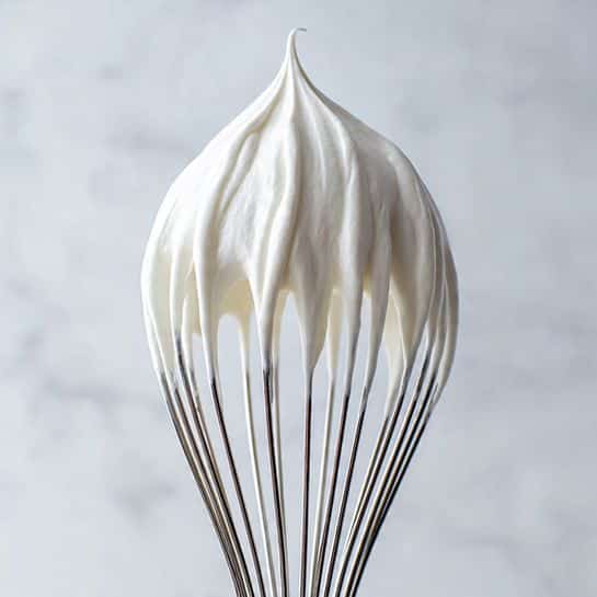 A close-up view of a dark metal whisk covered in smooth, glossy white whipped cream that peaks sharply at the top, with thick strands of cream stretching down between the whisk wires. The whisk sits on a white marbled surface, with the cream catching soft light that highlights its silky texture and slight shine. photo taken with an iphone --ar 4:5 --v 7