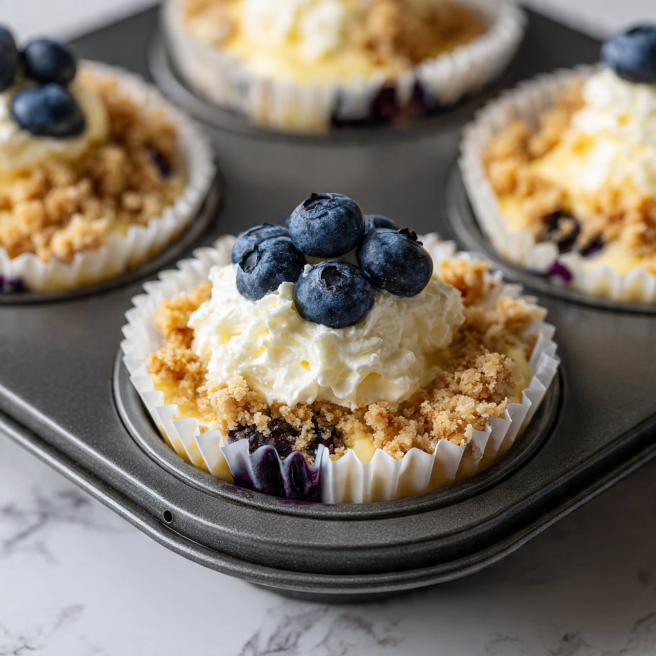 The image shows a close-up of a muffin cup lined with a white paper liner, sitting in a dark metal muffin tray on a white marbled surface. It has three layers visible: the bottom layer is a creamy pale yellow batter mixed with whole blueberries peeking through, giving spots of dark blue and shiny texture; the middle layer is a dollop of thick, white creamy mixture placed in the center on top of the batter and blueberries; the top layer consists of a crumbly golden brown streusel topping scattered with more whole fresh blueberries, adding a contrasting texture and dark blue color on the top. The layers combine to look smooth, creamy, soft, and crumbly all at once. Photo taken with an iphone --ar 4:5 --v 7