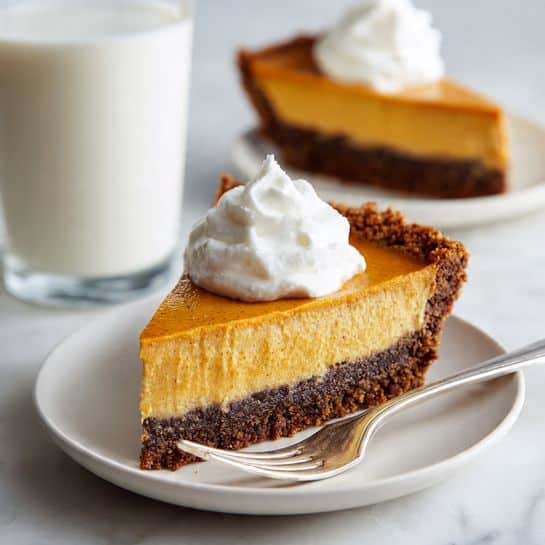 A slice of pie with three layers sits on a white plate with a silver fork. The bottom layer is dark brown, crumbly, and thick, forming a firm crust. Above it, there is a smooth orange layer that looks creamy and dense, taking up most of the slice. On top, a soft, white dollop of whipped cream sits in a small swirl. In the background, there is another plate with a similar pie slice and a tall glass of white milk, all placed on a white marbled surface. photo taken with an iphone --ar 4:5 --v 7