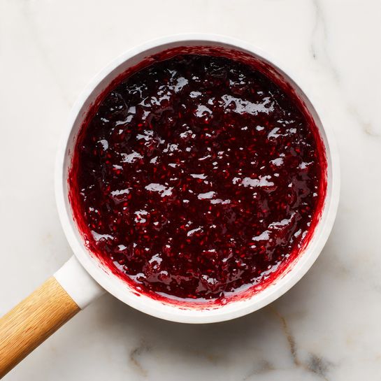 A close-up top view of a small white saucepan filled with thick, dark red, chunky sauce or jam. The sauce has a shiny and textured surface, showing bits of fruit within it. The saucepan has a light wooden handle and is placed on a white marbled surface, with some sauce slightly sticking to the inside edges of the pan. photo taken with an iphone --ar 4:5 --v 7