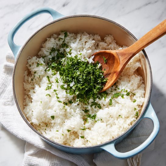 A light blue pot with two handles holds fluffy white rice that fills most of the pot. On top of the rice, a pile of finely chopped fresh green herbs is placed in the center, adding a bright contrast to the white rice. A wooden spoon is angled inside the pot, resting on the rice and herbs. The pot sits on a white marbled surface with a soft white cloth nearby. The scene is brightly lit, showing clear textures of the rice grains and fresh herbs, photo taken with an iphone --ar 4:5 --v 7