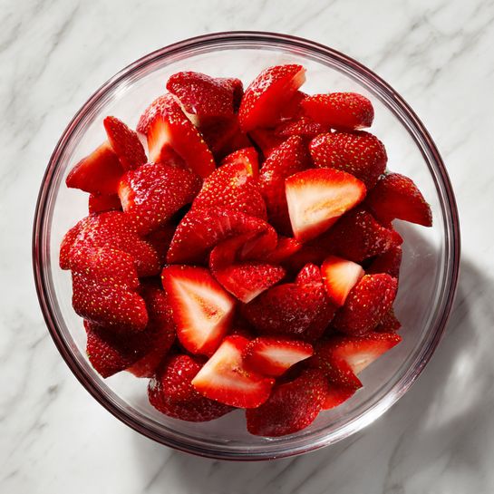 A clear glass bowl is filled with fresh red strawberries, each strawberry sliced into halves and quarters showing their bright, juicy inside. The strawberries have a natural shine, and some small seeds are visible on the outer red skin. The bowl sits on a white marbled surface. Photo taken with an iphone --ar 4:5 --v 7