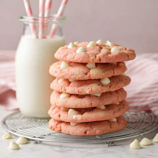 A stack of five round, soft pink cookies with a cracked surface is placed slightly off-center on a white marbled surface, each cookie topped with three or four small, smooth, pale yellow dollops. Some cookies lie flat on a metal cooling rack beside the stack, and a light pink fringed cloth is partially under the rack. A tall, white glass bottle stands blurred in the background, all set against a softly blurred white marbled background. photo taken with an iphone --ar 4:5 --v 7