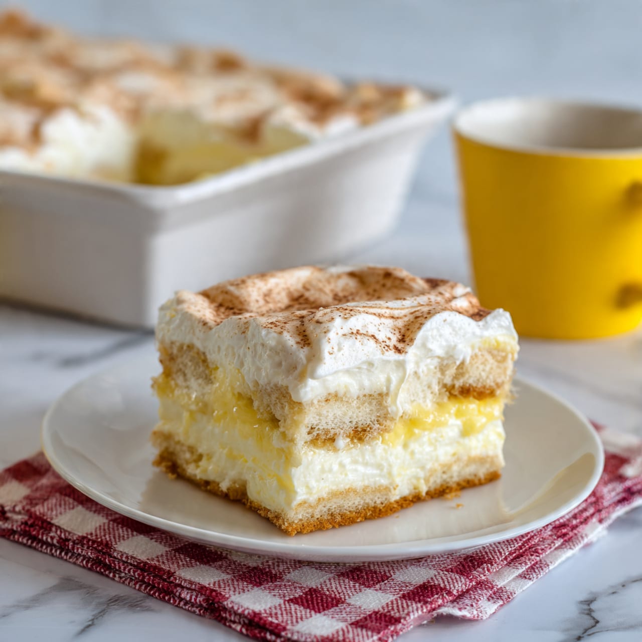 A square piece of layered dessert sits on a white plate with a red and white checkered cloth under it. The dessert has three visible layers: a light golden base layer with a soft texture, a thick creamy white middle layer that looks smooth and fluffy, and a top layer with a pale yellow creamy texture, slightly glossy with soft swirls, suggesting a light sauce or glaze. In the background, a white rectangular dish filled with more of the same dessert sits on a surface with a bright yellow cup beside it. The whole scene is set against a white marbled texture. Photo taken with an iphone --ar 4:5 --v 7