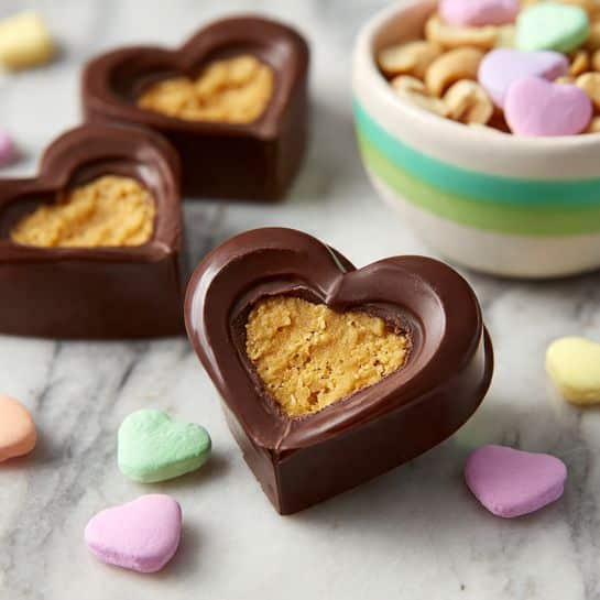 The image shows several heart-shaped chocolates with a smooth, shiny brown coating arranged on a white marbled surface. Each chocolate has a thick outer layer of chocolate, covering a dense, golden-brown filling visible in one open piece with a clean edge. The chocolates vary slightly in shape and size but all maintain the heart form. The surface of the chocolates has a polished texture with small bubbles and dimples that catch the light. photo taken with an iphone --ar 4:5 --v 7