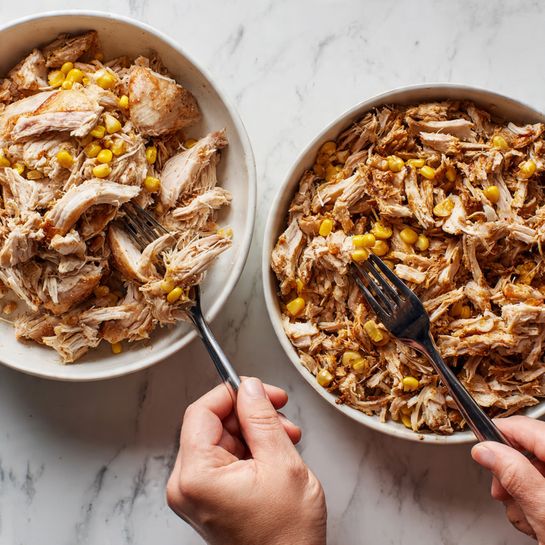 The image shows two white bowls on a white marbled surface with pieces of cooked chicken and some corn mixed in. The bowl on the left contains large, whole pieces of chicken with a light brown color and a few yellow corn kernels scattered on top. A woman's hand is using two black forks to pull apart one piece of chicken, revealing the tender, lighter inner meat. The bowl on the right has the chicken shredded into many smaller, uneven strips that are mixed with bits of corn, giving it a mix of brown with some yellow highlights. The texture of the chicken looks soft and moist in both bowls. photo taken with an iphone --ar 4:5 --v 7
