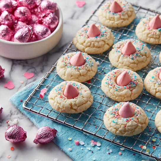 The image shows two types of round cookies on a metal cooling rack placed on a blue cloth with a white marbled surface underneath. One type of cookie is plain with a light beige cracked surface, and the other type is covered with colorful tiny round sprinkles. Each cookie has a pink chocolate kiss-like candy placed at the center, creating a pointed top. Scattered among the cookies are small shiny pink foil-wrapped chocolates. In the background, there is a white bowl filled with more pink foil-wrapped chocolates. The scene is bright and clean, focusing on the contrast between the cookies, sprinkles, and candies. Photo taken with an iphone --ar 4:5 --v 7