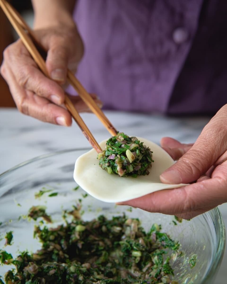 A woman's hand is holding a thin, round white dough piece with a small mound of green and light brown filling placed in the center by a pair of wooden chopsticks. The filling is made of finely chopped leafy greens mixed with small bits of other ingredients. Below the hand, a clear glass bowl is filled with more of the same green filling, resting on a white marbled surface. The person wearing a purple shirt is partially visible in the background, focusing on adding the filling onto the dough. The photo taken with an iphone --ar 4:5 --v 7