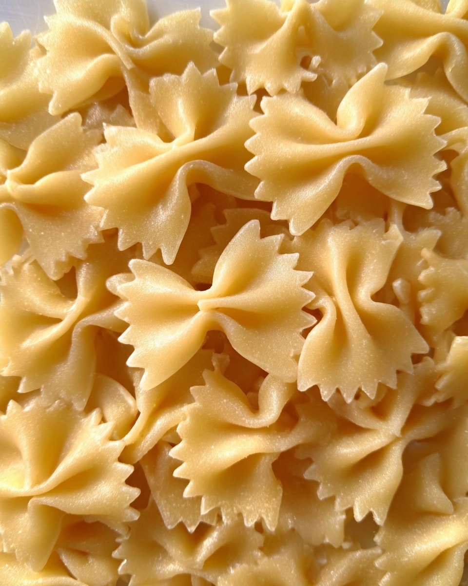 This image shows a close-up of cooked farfalle pasta. The bow-tie shaped pieces are pale yellow with smooth, slightly curved edges and ruffled sides. The pasta fills the entire frame, layered closely together with a soft texture that looks tender and lightly glossy. The background is a white marbled texture. Photo taken with an iphone --ar 4:5 --v 7
