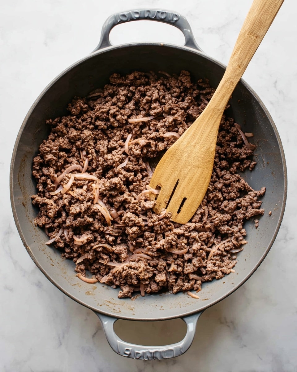 A large gray pan with two handles, filled with cooked ground meat that is brown and crumbly, shows some small pieces of onion mixed in. A wooden spatula lies inside the pan, partially resting on the meat, with a smooth texture and light brown color. The pan is placed on a white marbled surface. photo taken with an iphone --ar 4:5 --v 7