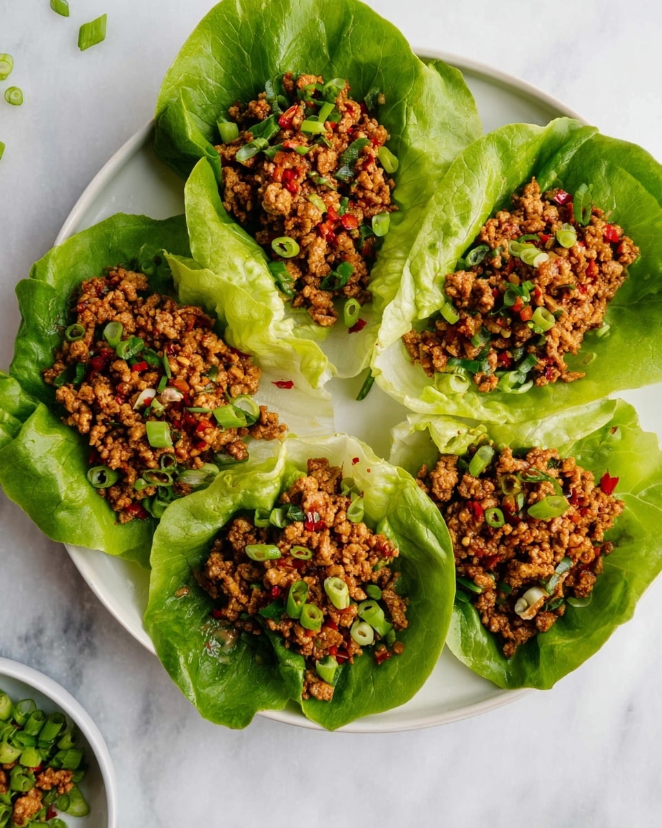 Five green lettuce leaves are arranged in a circle on a white plate, each leaf holding a layer of small bits of cooked brown meat mixed with chopped green onions and small red pieces, creating a textured and colorful topping. The lettuce leaves are fresh and crisp, with visible veins and slightly curled edges, and the meat mixture looks moist and well seasoned. Small chopped green onions are scattered on top of the meat and around the plate on the white marbled surface, giving a fresh garnish detail. Photo taken with an iphone --ar 4:5 --v 7