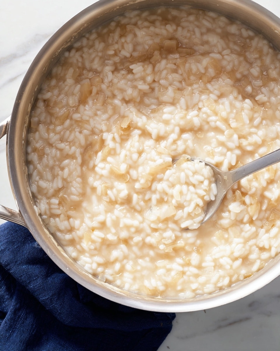 A close-up view of a silver cooking pot filled with creamy risotto, showing short, plump rice grains suspended in a thick, pale beige sauce with small bits of translucent onion mixed in. The risotto has a smooth, soft texture with a slight shine, and a stainless steel spoon is partially submerged in the mixture, stirring it gently. The pot sits on a white marbled surface with a dark blue cloth partially visible underneath it. photo taken with an iphone --ar 4:5 --v 7