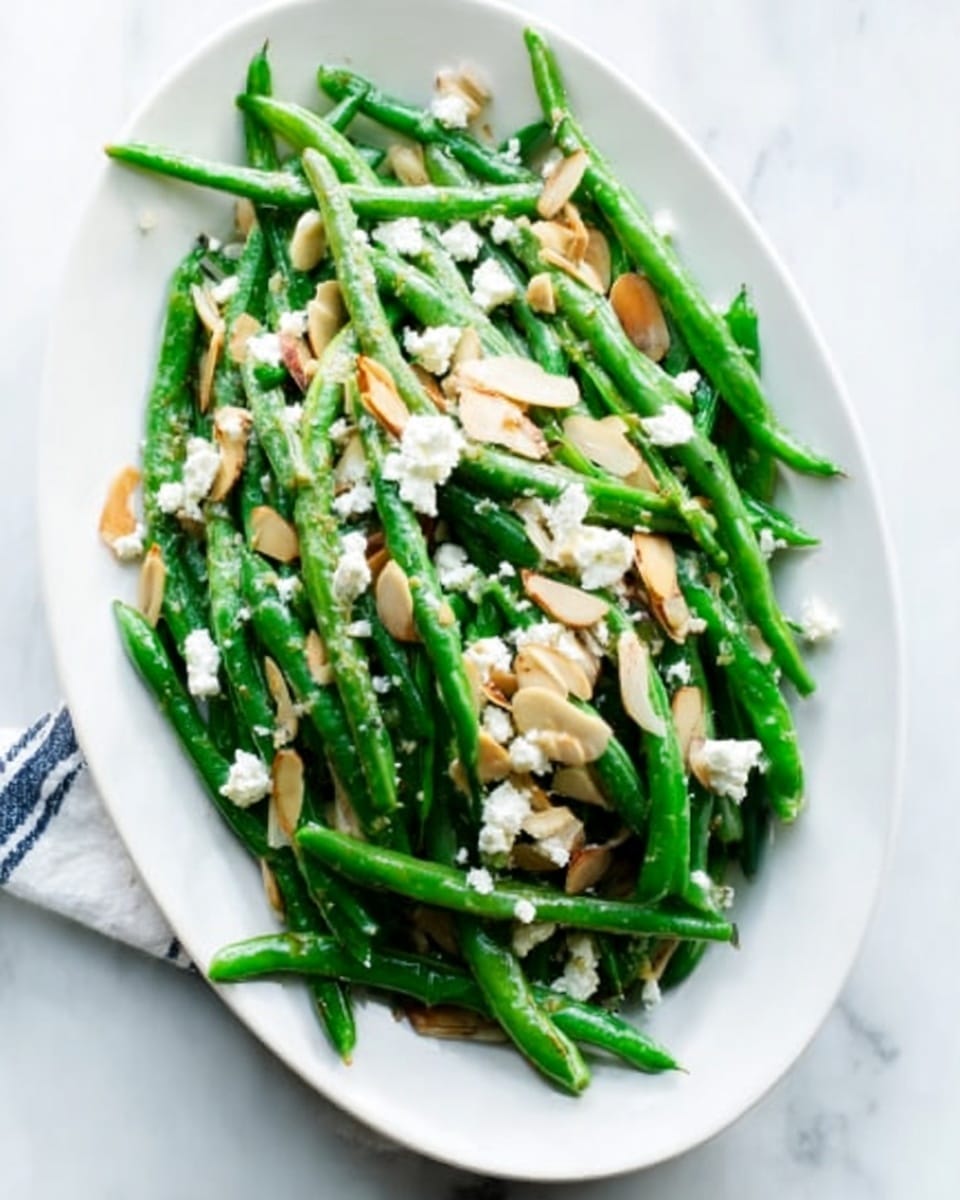 A white oval plate filled with green beans that are neatly arranged in a pile. On top of the green beans, there are small white crumbles, likely cheese, and thin, light brown slices of almonds scattered evenly. The plate is placed on a white marbled surface, adding a clean and fresh look to the scene. photo taken with an iphone --ar 4:5 --v 7