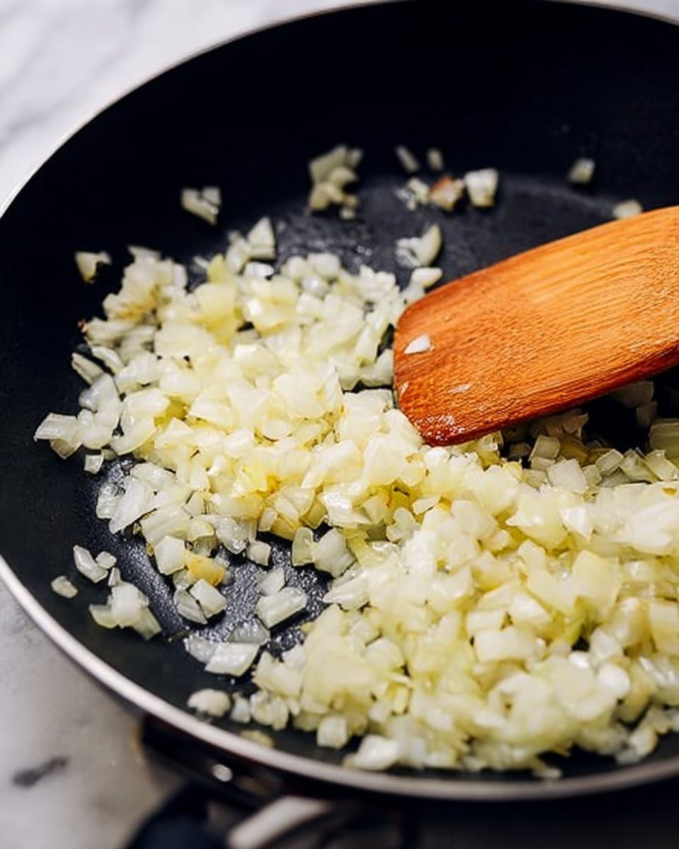 A close-up view of a black pan with a layer of small, light-yellow to white chopped onions spread evenly inside. A wooden spatula is positioned on the right side of the pan, touching the onions as if stirring them. The onions have a soft, slightly shiny texture showing they are cooking in some oil. The background features a white marbled surface. photo taken with an iphone --ar 4:5 --v 7