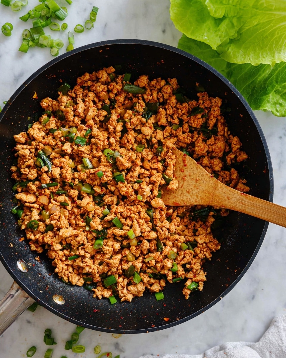 A black pan filled with cooked minced chicken mixed with small green pieces of chopped spring onions and light brown diced vegetables, all stirred together with a wooden spatula sitting inside the pan. The dish shows a mix of reddish-brown and pale yellow colors from the cooked chicken and seasoning, with green bits scattered throughout. The pan is on a white marbled texture surface, with a few chopped green onions scattered nearby and a few green lettuce leaves visible in the top left corner. Photo taken with an iphone --ar 4:5 --v 7