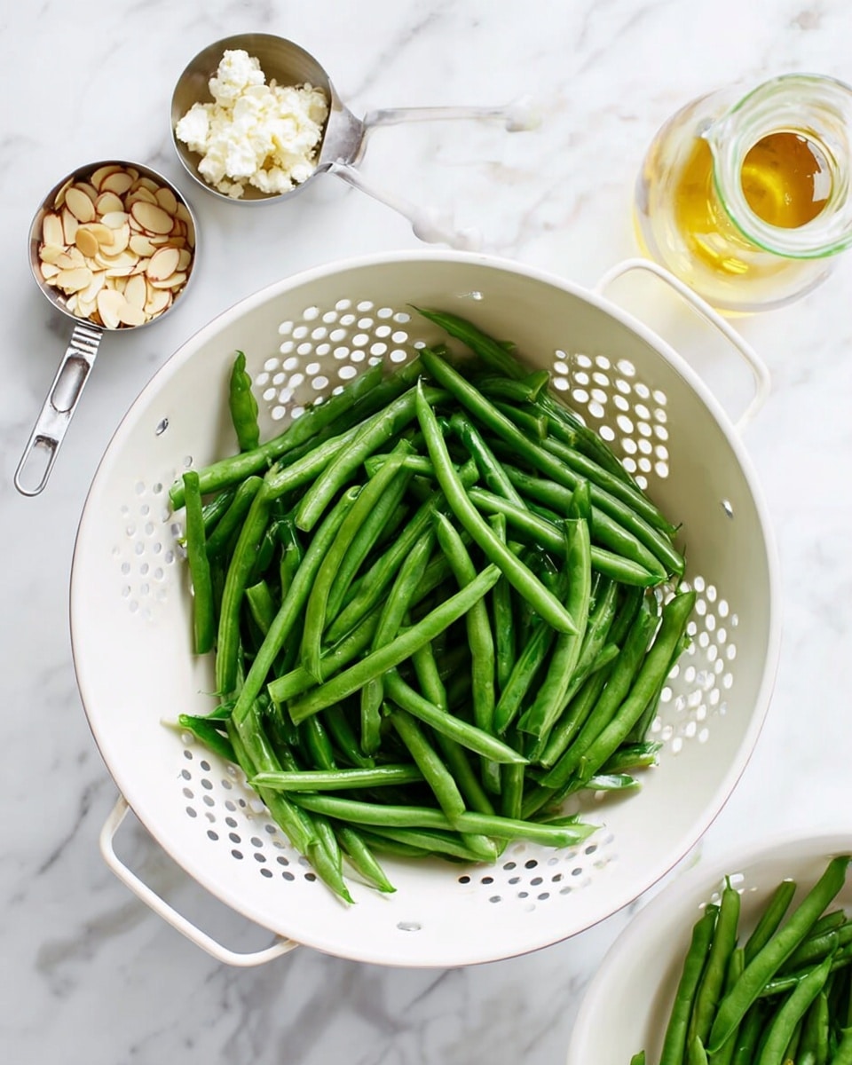 The image shows fresh green beans placed in a white colander with small holes, centered on a white marbled surface. Around the colander, there are three small metal measuring cups holding slivered almonds, crumbled cheese, and a clear glass bottle with a yellowish liquid, likely dressing or oil. The beans are bright green, smooth, and whole without any visible cuts or damage. The scene is bright and clean, with the white colander and utensils contrasting against the vibrant green beans. photo taken with an iphone --ar 4:5 --v 7