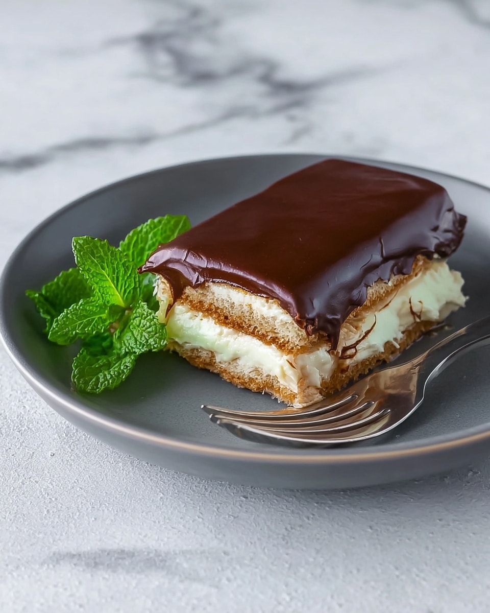 A slice of layered dessert is shown on a white plate, with three visible layers: the bottom and middle layers are light brown and textured like biscuit or cake, sandwiched with thick, smooth white cream layers in between. The top layer is rich dark brown chocolate that looks smooth and thick, evenly spread over the dessert. To the left side of the dessert on the plate, there is a small bunch of fresh green mint leaves adding color contrast. A silver fork rests beside the dessert on the plate. The background is a white marbled surface with a blue and white checkered cloth partially visible behind the plate. Photo taken with an iphone --ar 4:5 --v 7