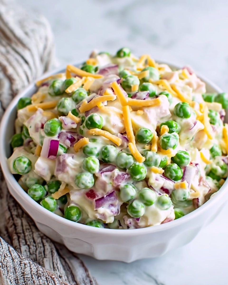 A white bowl filled with a creamy salad showing three main layers: bright green peas scattered throughout, small pieces of white onion and red onion adding specks of purple and white, and thin yellow strips of cheese mixed evenly. The salad has a smooth creamy texture coating all ingredients, creating a soft shine. The bowl sits on a white marbled surface in soft daylight. Photo taken with an iphone --ar 4:5 --v 7