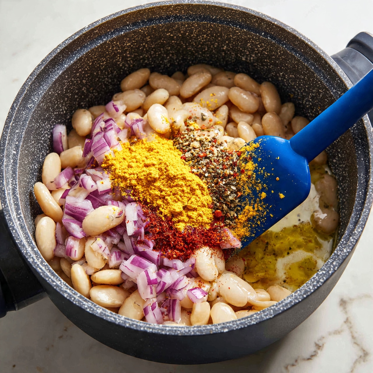 The image shows a close-up view inside a dark gray cooking pot with a speckled texture. Inside, there are white beans mixed with small pieces of purple onions. On top of the beans, there are several bright yellow, red, and black spices piled in the center. A blue spatula is scooping the spices, partially covered in the yellow powder. The liquid in the pot is creamy white with yellow oil droplets floating on the surface. The white marbled surface underneath the pot is visible around the edges. photo taken with an iphone --ar 4:5 --v 7