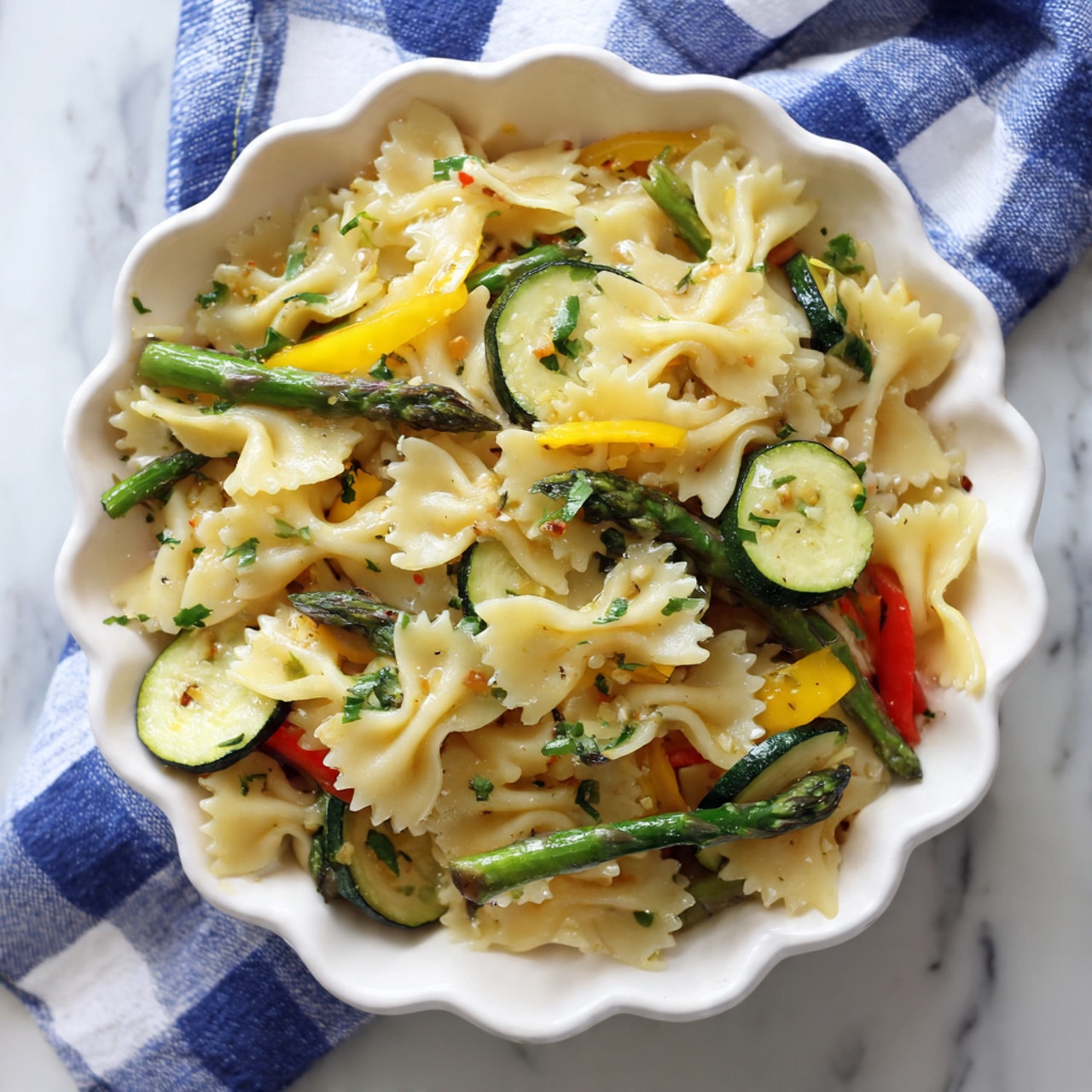 The image shows a white scalloped bowl filled with farfalle pasta mixed with bright green asparagus, sliced zucchini, yellow squash, and small pieces of yellow bell pepper. The pasta is off-white and soft-looking, while the vegetables add vibrant green and yellow colors with varied textures like the smooth zucchini slices and the firm asparagus tips. The bowl is placed on a white marbled surface with a blue and white checkered cloth partially underneath. Photo taken with an iphone --ar 4:5 --v 7