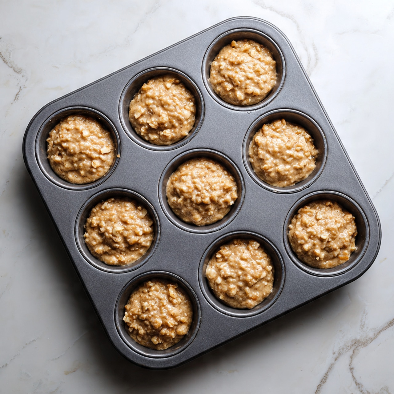 A dark gray metal muffin tray with twelve round cups, each filled nearly to the top with light brown, uneven textured batter. The batter looks moist and thick with small chunks visible, giving a rough and slightly lumpy surface in each cup. The tray is placed on a white marbled surface with faint gray veins, and soft natural light highlights the batter’s texture, showing a mix of smooth and coarse parts within each cup. photo taken with an iphone --ar 4:5 --v 7