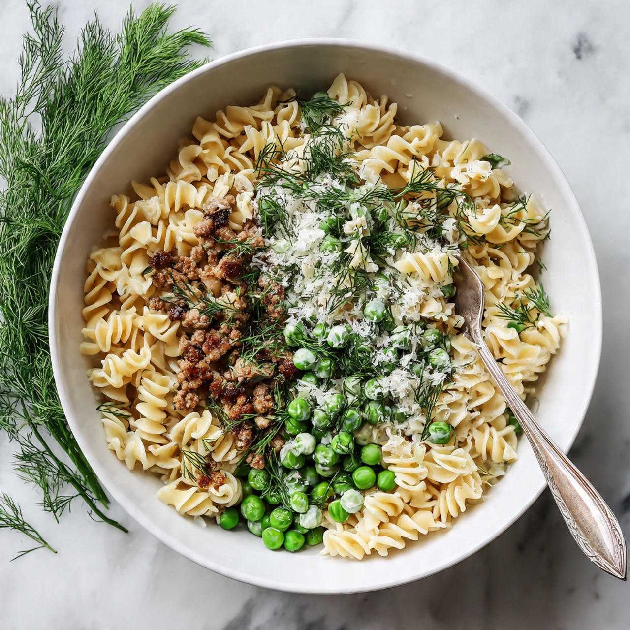 A white bowl filled with three layers of food: the bottom layer is wavy-edged pasta with a light creamy color and soft texture, the middle layer shows small green peas and chopped green herbs mixed with browned crumbled meat pieces, and the top layer is sprinkled with white grated cheese and fresh green dill leaves. A silver fork is resting inside the bowl, its handle touching the edge and tines partially buried under the pasta. The bowl is placed on a white marbled surface with some green dill sprigs beside it. Photo taken with an iphone --ar 4:5 --v 7