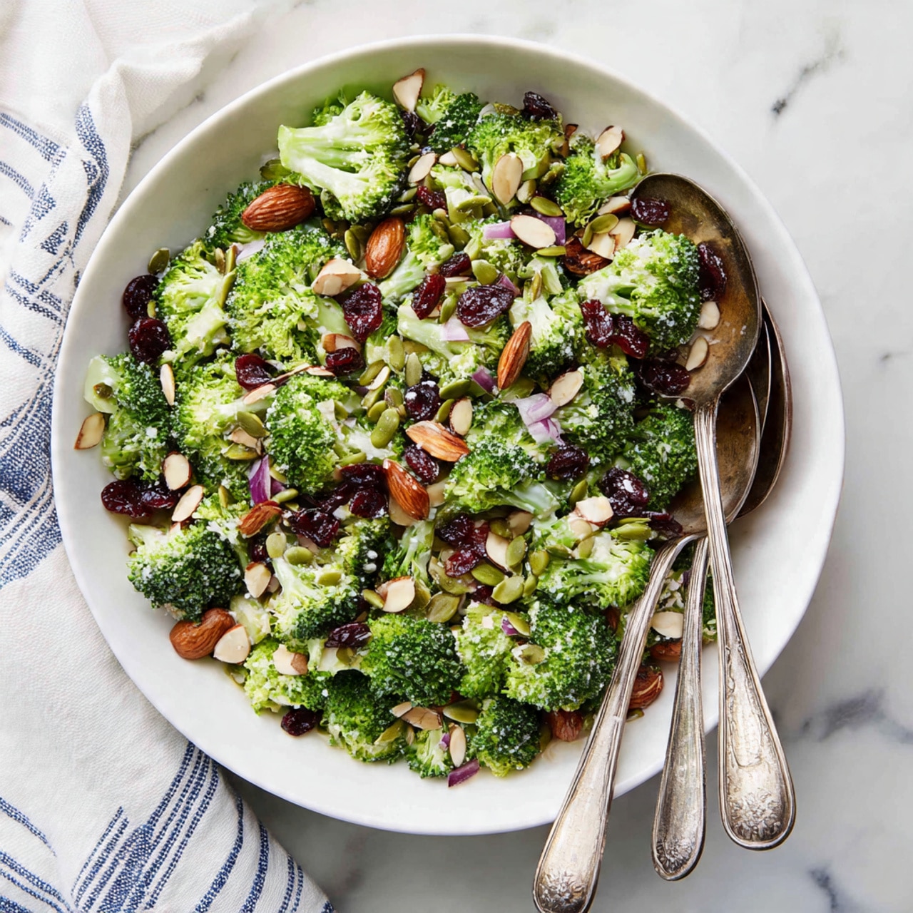 A white round plate filled with a fresh broccoli salad showing many small, green broccoli florets as the main layer, mixed with scattered red dried cranberries adding pops of dark red color, and topped with whole brown almonds and flat green pumpkin seeds spread evenly across. Small pieces of white and purple diced onion are sprinkled throughout, adding texture and color contrast. Two vintage silver spoons rest on the right side of the plate, partially touching the salad, all set on a white marbled surface with a white and blue striped cloth nearby. Photo taken with an iphone --ar 4:5 --v 7
