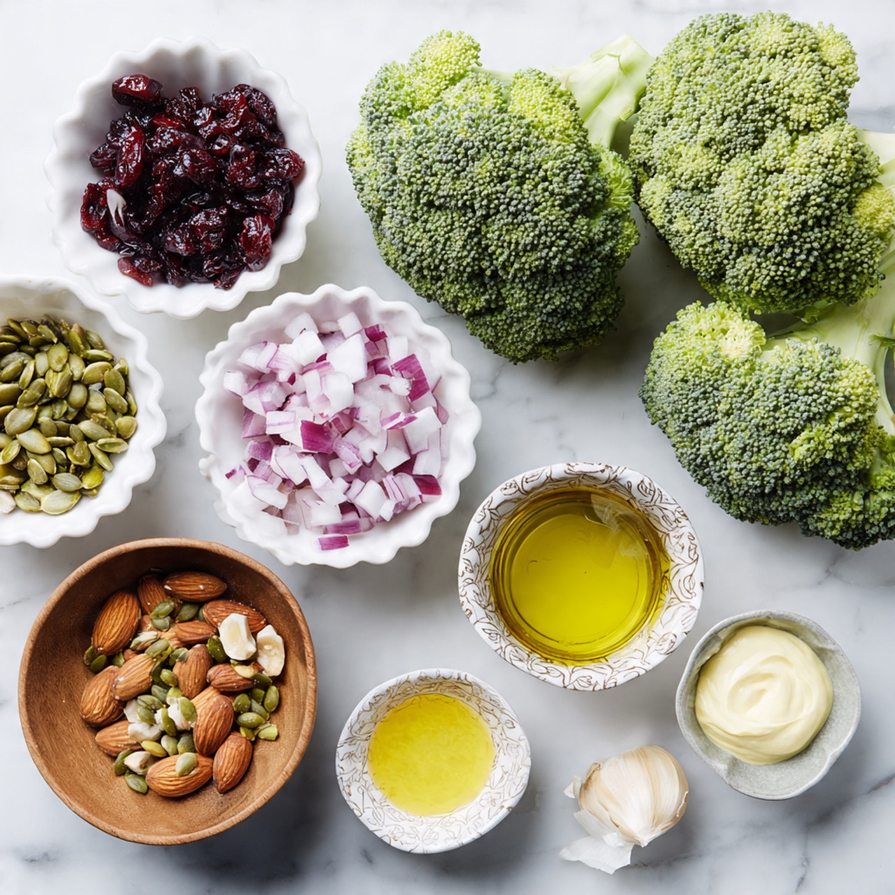 The image shows a group of ingredients on a white marbled textured surface. At the top, there are two green broccoli heads with textured florets and thick light green stems. Below and to the left, a small white bowl holds dark red dried cranberries, and next to it is a white scalloped bowl filled with small diced purple and white onion pieces. Further down on the left side, a wooden bowl contains a mix of toasted almonds and green pumpkin seeds. To the right of that, four small white or patterned bowls are arranged, holding light yellow creamy mustard, golden olive oil in a glass container, amber-colored vinegar in a patterned bowl, and pale creamy mayonnaise. A small piece of garlic clove is placed near the top right corner of the bowls. photo taken with an iphone --ar 4:5 --v 7