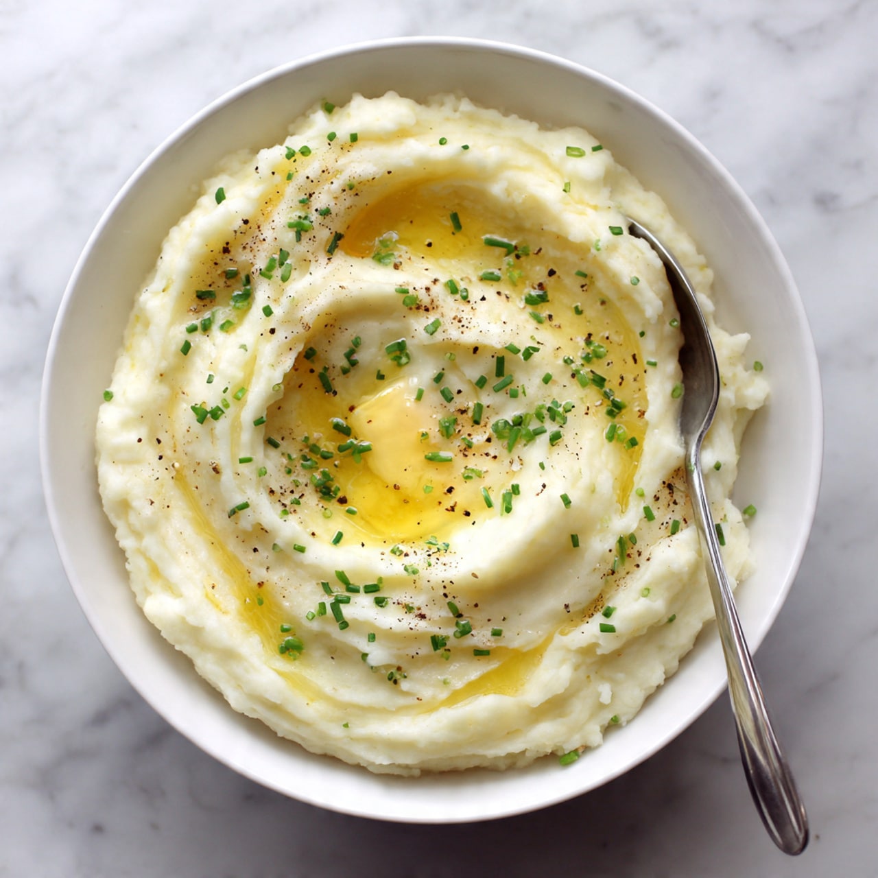 A white bowl filled with a smooth, creamy layer of mashed potatoes with light yellow butter melting in the center. The potatoes have a soft texture with gentle swirls and a few small peaks. The top layer is sprinkled with small green chives scattered evenly and a light dusting of black pepper. A silver spoon is resting inside the bowl on the right side, slightly sinking into the mashed potatoes. The bowl sits on a white marbled surface. photo taken with an iphone --ar 4:5 --v 7