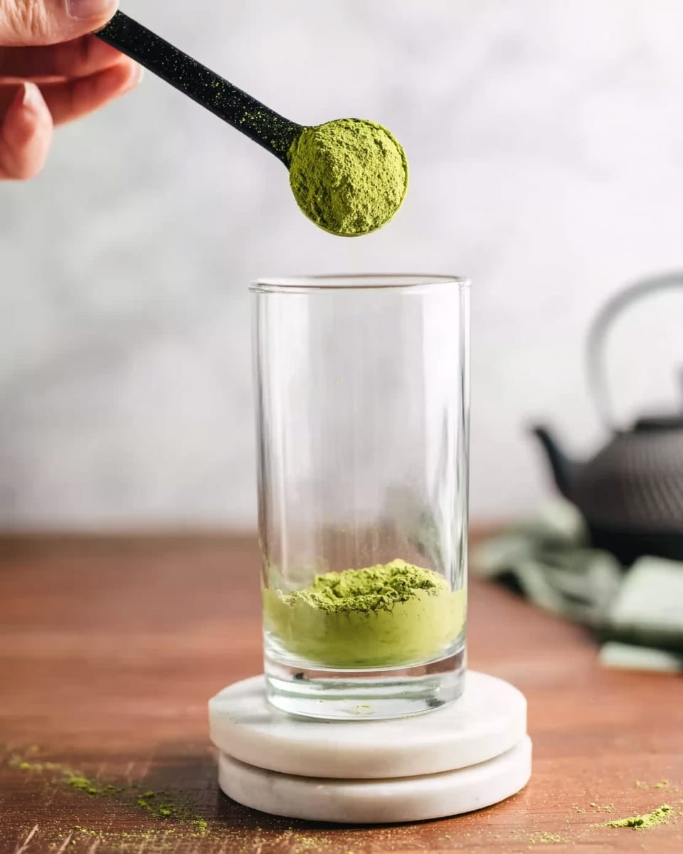 A tall clear glass is shown with one small heap of bright green powder resting at the bottom inside the glass. Above it, a woman's hand holds a black measuring spoon filled with the same bright green powder, about to drop it into the glass. The glass sits on two stacked white marble coasters on a wooden table. The background and surface are white marbled texture with blurred kitchen items including a teapot. Photo taken with an iphone --ar 4:5 --v 7