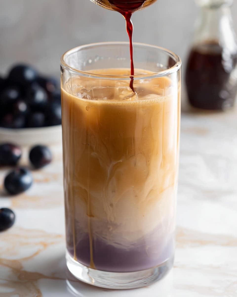 A clear glass filled with a three-layer iced drink stands on a white marbled surface. The bottom layer is light purple with a smooth, slightly swirling texture. The middle layer is a soft light brown blending gently into the bottom. The top layer is a creamy caramel color, smooth and thick, almost like a soft foam. Thin dark red liquid is being poured into the glass from above, creating small ripples on the caramel layer. In the background, there are blurred dark blue berries and a small glass bottle. Photo taken with an iphone --ar 4:5 --v 7