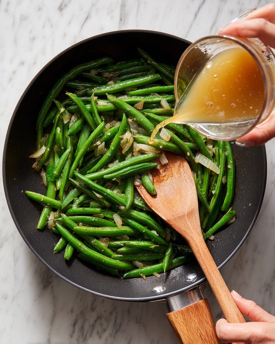 A black frying pan with a wooden handle sits on a white marbled surface. Inside the pan is a layer of cooked green beans mixed with small pieces of onion, both slightly shiny from cooking. A wooden spoon is resting inside the pan, touching the green beans. A woman's hand is pouring a light brown liquid from a clear measuring cup into the pan. The scene shows mid-action cooking with fresh green beans and onions in the pan. Photo taken with an iphone --ar 4:5 --v 7