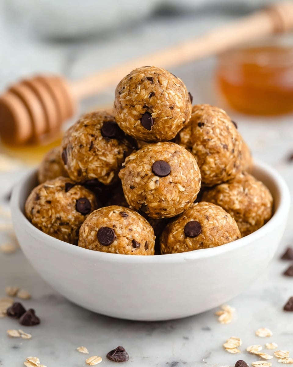 A white bowl filled with round, golden-brown energy balls that have a rough texture from oats and small dark chocolate chips scattered throughout each ball. The balls are stacked in layers inside the bowl, with some chips visibly embedded on the surface, giving a slightly bumpy look. In the background, there is a blurred honey dipper with honey and a white marbled surface beneath the bowl. Some scattered oats and chocolate chips lie near the bowl. Photo taken with an iphone --ar 4:5 --v 7