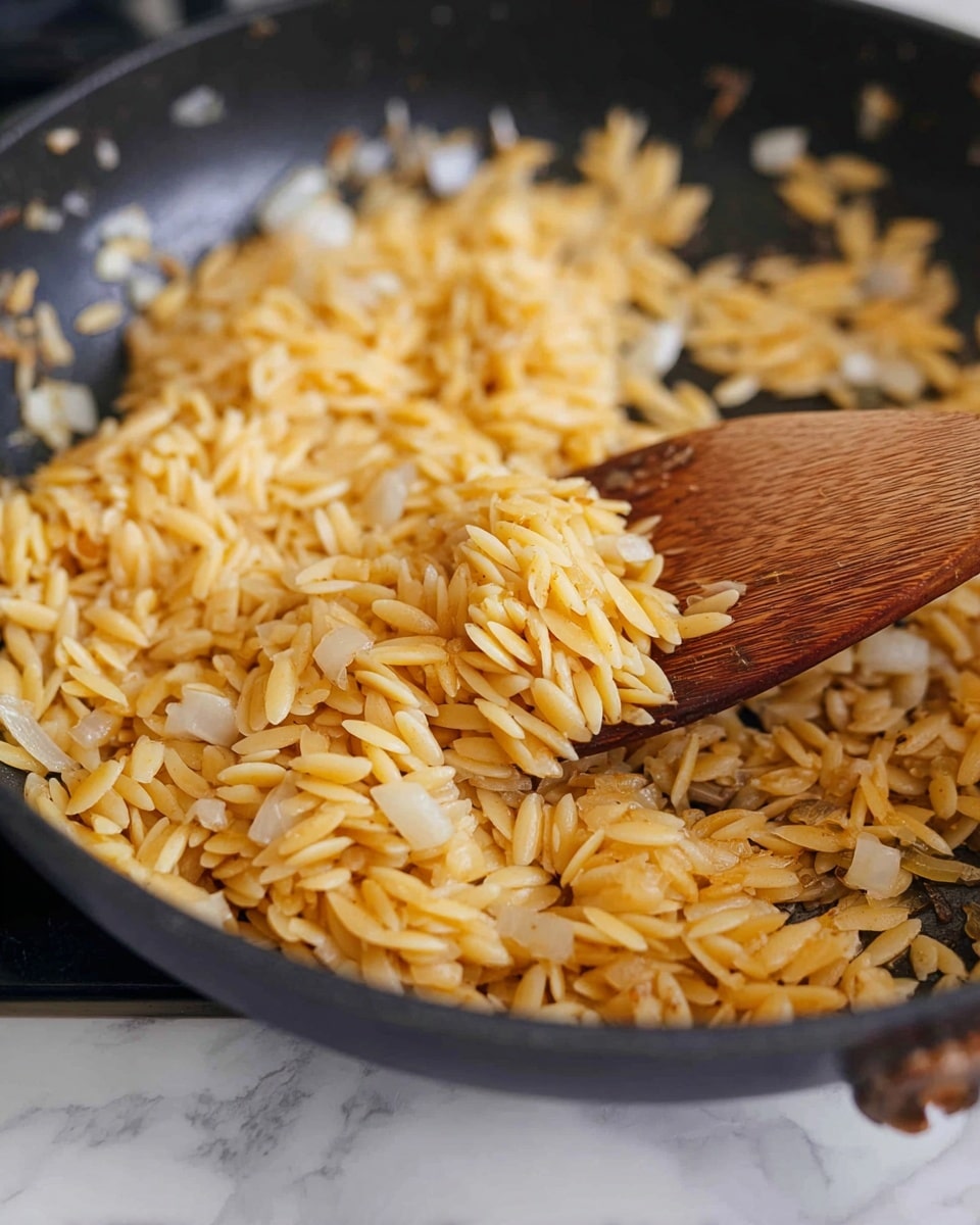 A close-up of a black pan filled with a single layer of golden toasted orzo pasta mixed with cooked small pieces of translucent white onion. A wooden spoon stirs the orzo and onion, showing the texture of the toasted pasta with a slight shine from oil. The background is a white marbled texture with soft, blurred shapes. Photo taken with an iphone --ar 4:5 --v 7