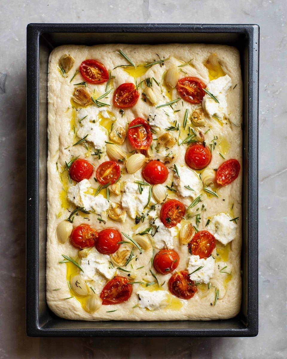 A rectangular black baking pan filled with one thick layer of unbaked light beige dough, topped with scattered bright red cherry tomatoes, whole golden brown garlic cloves, and uneven dollops of white soft cheese, all drizzled with yellow olive oil and sprinkled with small green rosemary leaves. The pan is placed on a white marbled texture surface. Photo taken with an iphone --ar 4:5 --v 7