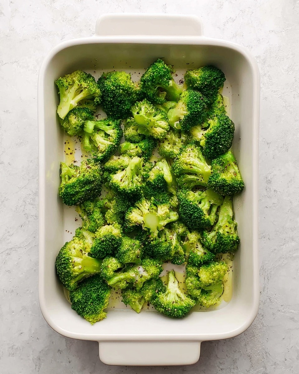 A white rectangular baking dish contains a single layer of bright green broccoli florets scattered evenly across the bottom. The florets have a fresh texture with tiny detailed buds, and spots of black pepper and a light drizzle of olive oil add a slight shine and contrast. The dish sits on a white marbled surface, creating a clean and fresh setting. Photo taken with an iphone --ar 4:5 --v 7
