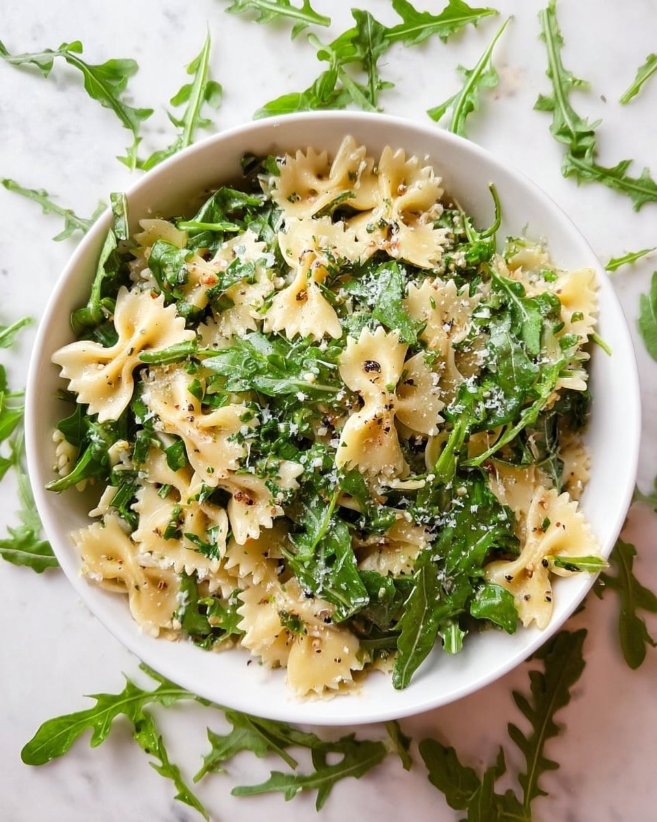 A white bowl filled with farfalle pasta mixed with fresh, bright green arugula leaves, scattered evenly throughout. The pasta is light yellow, with a slightly glossy texture from a light dressing, and small bits of grated cheese sprinkled on top, adding a fine white dusting. The arugula leaves have delicate veins and a crisp texture, giving the dish a fresh look. Around the bowl, loose arugula leaves are scattered on a white marbled surface. photo taken with an iphone --ar 4:5 --v 7