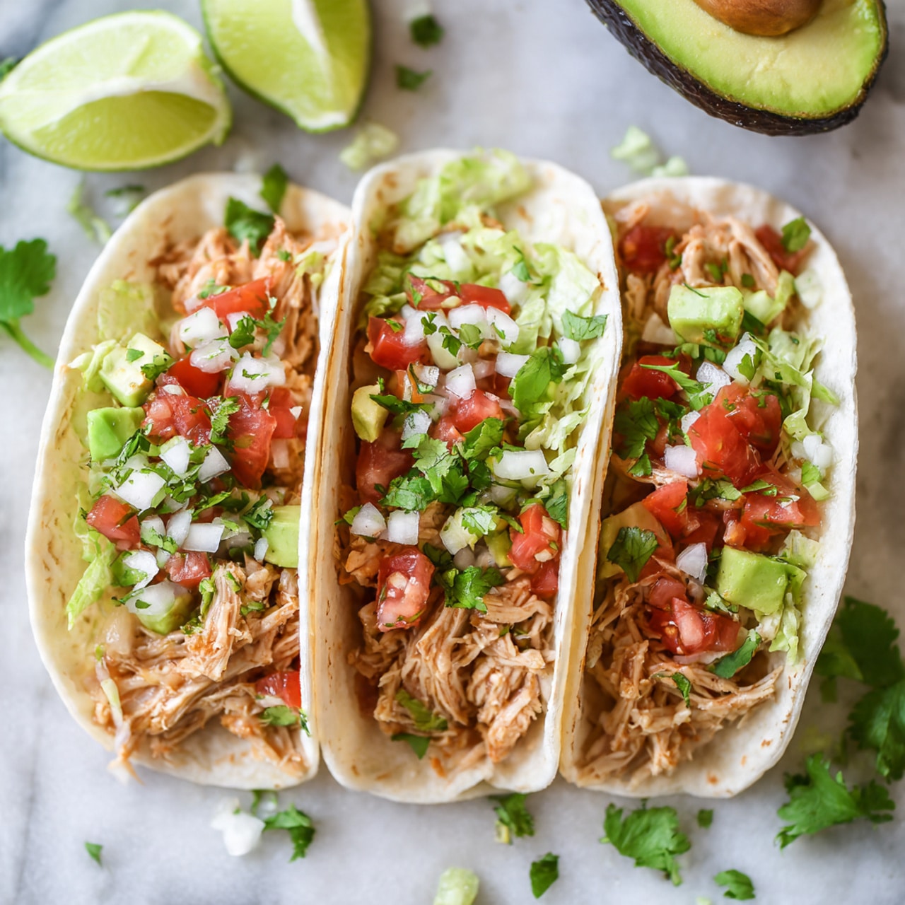 Two soft, white tortillas form the base, each folded slightly to hold the fillings. The first layer inside is shredded light-brown cooked chicken. On top of the chicken, there is a mix of finely chopped red tomatoes, white onions, and green cilantro creating a fresh pico de gallo. Chopped green avocado cubes and shredded pale green lettuce add more color and texture on each taco. The tacos rest on a white marbled textured surface with a few lime wedges and part of an avocado visible in the background. The overall look is fresh, colorful, and vibrant. photo taken with an iphone --ar 4:5 --v 7